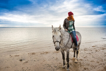 Fototapeta na wymiar wearing jacket, jeans and warm hat female horse jokey goes on horseback along coast