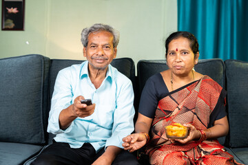 Senior couple watching tv or television at home by surfing channels - concept of relaxation,...