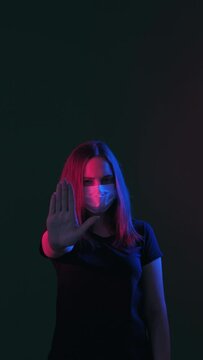 Stop sign. Stay home. Woman in protective mask neon light glow showing forbidden hand gesture looking at camera isolated on black vertical. Quarantine measures. Covid-19 restriction. New normal