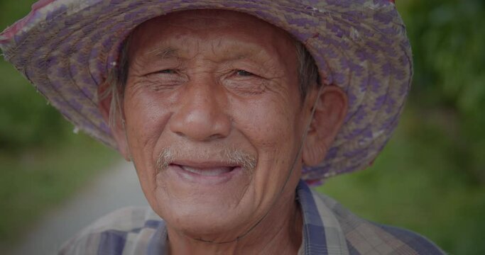 Slow motion scene of happy smiling and laughing Asian man who is poor elderly villager, older than seventy stands wearing old hat leading the bicycle on the roadside of his village.