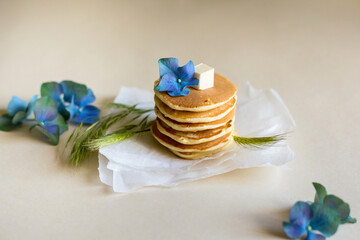 Foodphoto. On a beige background - pancakes on a paper backing. Decorated with oil, blue hydrangea flowers, green spikelets. Nice breakfast and lunch. Daylight. Illustration for a recipe.