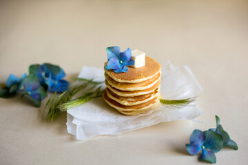 Foodphoto. On a beige background - pancakes on a paper backing. Decorated with oil, blue hydrangea flowers, green spikelets. Nice breakfast and lunch. Daylight. Illustration for a recipe.