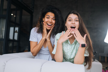 Two girls sit on the back of the sofa, look at the cameras and smile