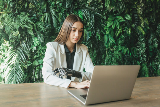 A beautiful young girl with a prosthetic bionic arm is studying or working on a laptop against a wall of plants. Online work, distance learning