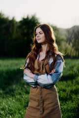 A farmer woman in her work clothes, plaid shirt and apron, stands in the field on the green grass and smiles in the setting sun