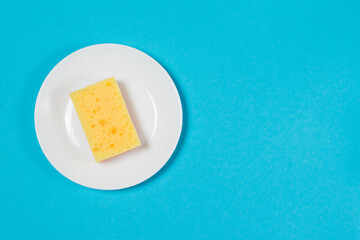 Yellow porous sponge for washing dishes on a white plate on a blue background with copy space. The concept of cleaning, washing dishes and doing housework.