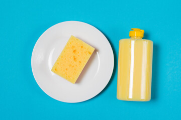 Yellow porous sponge for washing dishes on a white plate and detergent in a plastic transparent bottle on a blue background with copy space. Concept of cleaning, washing dishes and doing housework.