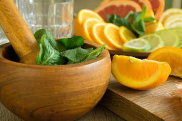Close-up of wooden mortar and pestle with leaves of green mint on colorful background of slices fresh sliced citrus fruits and glass. Lobules of orange, lemon, lime, grapefruit lying on cutting board