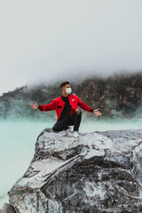 young man in red bomber jacket kneeling on rock at kawah putih sulfer lake