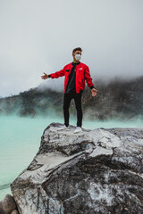 young man in red bomber jacket standing on rock at kawah putih sulfer lake