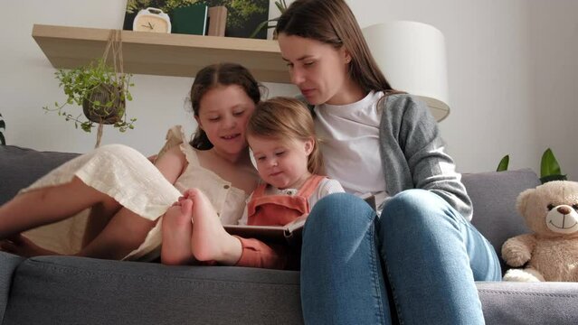 Happy young mother babysitter hold read book or photo album relax embrace cute little children daughters, smiling mum tell small kids funny fairy tale story sitting on sofa having fun together at home