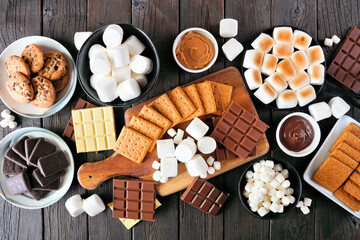 Smores buffet table scene. Roasted marshmallows, crackers, chocolate and a variety of ingredients. Overhead view on a dark wood background.
