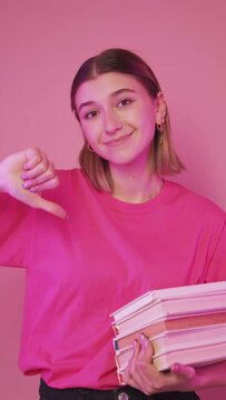Bad book. Study pressure. Exam deadline. Rejecting disappointed woman student with pile of homework textbooks showing thumb down gesture isolated on pink vertical.