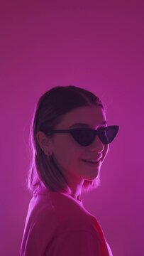 Sunglasses fashion. Cool woman. Trendy accessory. Confident stylish pretty girl winking eye in vibrant neon light isolated on pink background vertical.