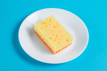 Sponge for washing dishes and cleaning in the apartment on a plate on a blue background. The concept of using environmentally friendly detergents for the home