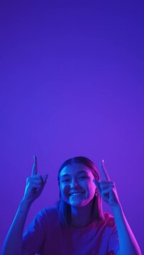 Pointing up girl. Discount offer. Promotional background. Joyful young woman advertising empty space purple ad in bright neon light vertical.