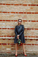 Portrait of young woman wearing black dress, leather jacket, holding bouquet of red roses, standing at brick wall.