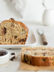 Banana bread with dark chocolate. Incision and piece of banana cake on the perchament paper. Cake decorated bananas on the white ceramic rectangular plate. High key. Table-knife with wooden handle