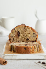 Banana bread with dark chocolate. Incision and piece of banana cake on the perchament paper. Cake decorated bananas on the white wooden serving board. High key