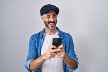 Hispanic man with beard using smartphone typing message winking looking at the camera with sexy...