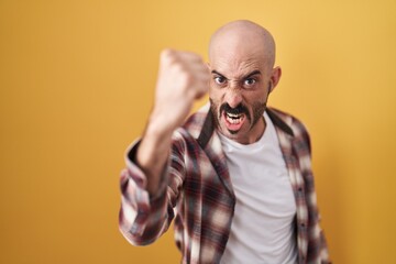 Hispanic man with beard standing over yellow background angry and mad raising fist frustrated and furious while shouting with anger. rage and aggressive concept.