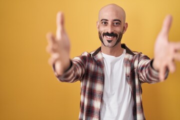 Fototapeta premium Hispanic man with beard standing over yellow background looking at the camera smiling with open arms for hug. cheerful expression embracing happiness.