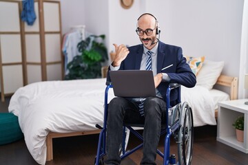 Hispanic man with beard sitting on wheelchair doing business video call pointing thumb up to the side smiling happy with open mouth