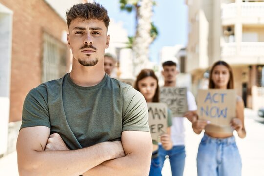 Young hispanic activist man with arms crossed gesture standing with a group of protesters holding banner protesting at the city.