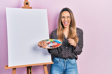 Beautiful hispanic woman standing drawing with palette by painter easel stand winking looking at...