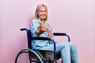 Beautiful blonde woman sitting on wheelchair beckoning come here gesture with hand inviting welcoming happy and smiling