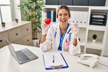 Young blonde woman wearing doctor uniform holding blood and heart smiling and laughing hard out...