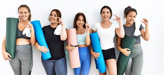 Obraz premium Group of women holding yoga mat standing over isolated background smiling and confident gesturing with hand doing small size sign with fingers looking and the camera. measure concept.