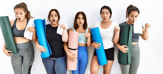 Group of women holding yoga mat standing over isolated background pointing down with fingers showing advertisement, surprised face and open mouth