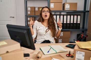 Young hispanic woman working at small business ecommerce amazed and surprised looking up and pointing with fingers and raised arms.