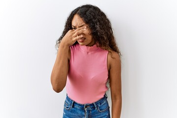 Beautiful hispanic woman standing over isolated background smelling something stinky and...