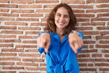 Beautiful brunette woman standing over bricks wall pointing to you and the camera with fingers, smiling positive and cheerful