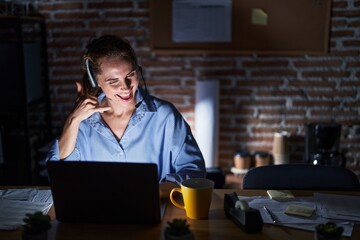 Beautiful brunette woman working at the office at night smiling doing phone gesture with hand and fingers like talking on the telephone. communicating concepts.
