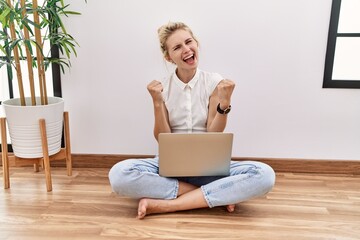 Young blonde woman using computer laptop sitting on the floor at the living room very happy and excited doing winner gesture with arms raised, smiling and screaming for success. celebration concept.