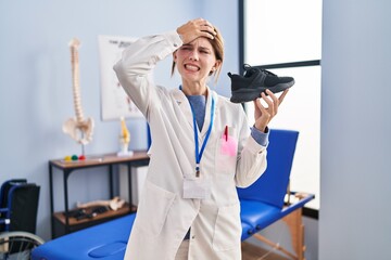 Young physiotherapist woman holding sneakers stressed and frustrated with hand on head, surprised and angry face