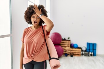 African american woman with afro hair holding yoga mat at pilates room surprised with hand on head...