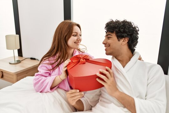 Young couple smiling happy holding valentine heart box gift at bedroom.