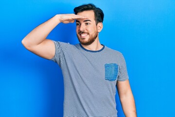 Handsome caucasian man with beard wearing casual striped t shirt very happy and smiling looking far away with hand over head. searching concept.