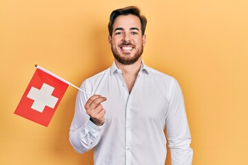 Handsome caucasian man with beard holding switzerland flag looking positive and happy standing and...