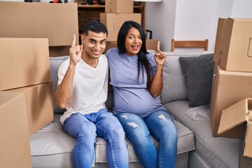 Young hispanic couple expecting a baby sitting on the sofa at new home smiling with an idea or question pointing finger with happy face, number one