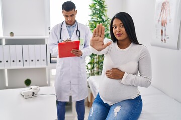 Fototapeta premium Young hispanic mother expecting a baby at the doctor with open hand doing stop sign with serious and confident expression, defense gesture