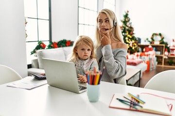 Mother of caucasian daughter wearing call center agent headset at home serious face thinking about question with hand on chin, thoughtful about confusing idea
