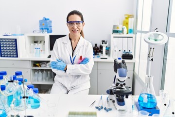 Young hispanic woman wearing scientist uniform with arms crossed gesture at laboratory
