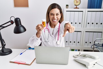 Young hispanic woman wearing doctor uniform holding pills at the clinic pointing to you and the camera with fingers, smiling positive and cheerful