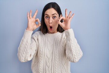 Young brunette woman standing over blue background looking surprised and shocked doing ok approval symbol with fingers. crazy expression