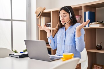 Young brunette woman wearing call center agent headset clueless and confused expression with arms...